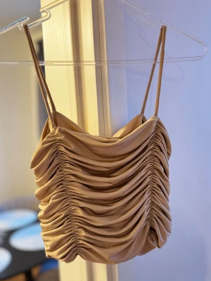 Beige/guld crop top - Trendig beige/guldig crop top med tunna axelband och snygga draperingar både fram och bak. Toppen har en glansig finish och är gjord i ett stretchigt material som sitter tajt. Perfekt för dig som vill ha en cool och modern look. Klippt bort lappen men är ganska säker att toppen är köpt på newyorker för ett x antal år sedan. Passar xs/s