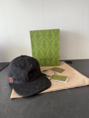Svart keps från Gucci med GG-mönster - Snygg svart keps från Gucci med klassiskt GG-monogram över hela kepsen och en grön-röd rand på sidan. Kepsen har böjd skärm och justerbar rem bak. Tillverkad i canvasmaterial och levereras med originalförpackning och tags.