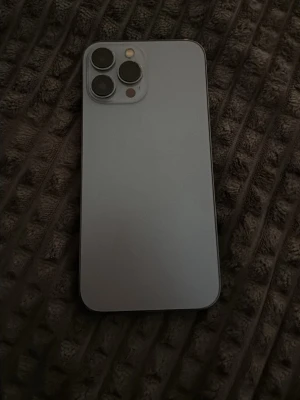 Silverfärgad iPhone med trippelkamera - Snygg iPhone i silver med stilren design och platt baksida i glas. Telefonen har tre stora kameralinser och en LED-blixt på baksidan. Skärmen är stor och har en tydlig notch upptill. Perfekt för dig som gillar Apple och vill ha en modern smartphone. iPhone 13 Pro Max. Batterihälsa 80%