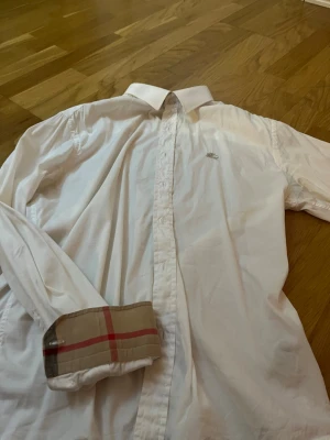 Vit skjorta från Burberry med logga - Klassisk vit skjorta från Burberry med diskret broderad logga på bröstet och ikoniskt rutigt tyg på insidan av manschetterna. Skjortan har knappar framtill och lång ärm. Perfekt för dig som gillar stilrena plagg med exklusiva detaljer.