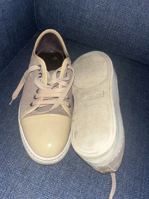 Beige sneakers från Lanvin - Säljer ett par beige sneakers från Lanvin med rund tå och gummitå. Skorna har beige textilovandel, matchande snören och vit sula med Lanvin-logga undertill. Snygg och stilren design som passar till det mesta.