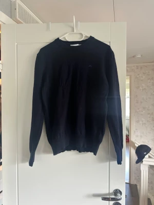 Mörkblå stickad tröja J.Lindeberg - Snygg mörkblå stickad tröja från J.Lindeberg med rund halsringning och diskret logga på bröstet. Tröjan har långa ärmar och ribbade muddar vid ärmslut och nederkant. Perfekt att matcha med jeans eller chinos.