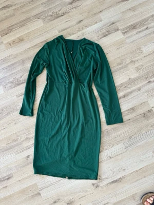 Grön omlottklänning med v-ringning - Stilren grön klänning med omlott och djup v-ringning. Långärmad modell i mjukt material, perfekt för dig som gillar enkel men snygg design. Klänningen har en rak passform och går ner till knäna.