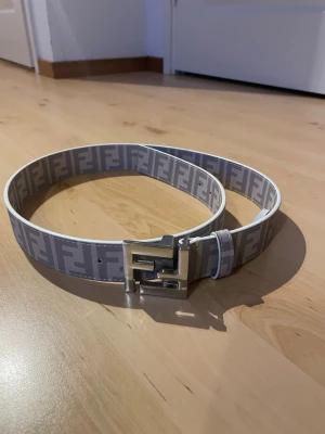 Gråvitt Fendi bälte med FF-logga - Säljer ett gråvitt bälte från Fendi med det klassiska FF-mönstret längs hela bandet och en stor metallspänne med Fendi-logga. Bältet har en modern och clean look som sticker ut och passar till många olika outfits.