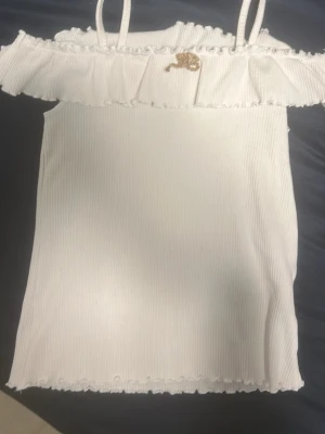 Vit off shoulder topp med volang - Trendig vit ribbad topp med smala axelband och volangdetalj runt axlarna. Off shoulder-modell med liten guldfärgad dekoration framtill. Perfekt för en somrig och fräsch look.