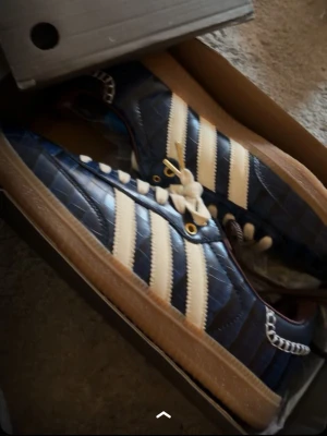 Adidas x walesbonner  - Säljer ett par blå Adidas sneakers med vit kontrastsöm och de klassiska tre vita ränderna på sidan. Skorna har en unik krokodil-liknande textur på ovandelen, vita skosnören och gummisula i brunt. Gulddetaljer vid snörningen och snygga detaljer runt öppningen.