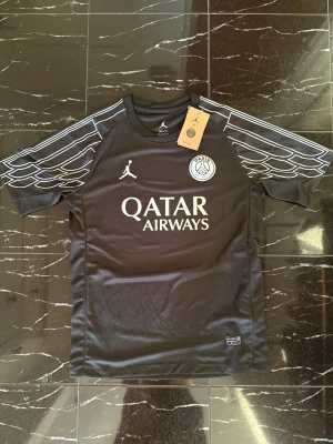 Svart PSG x Jordan matchtröja - Snygg svart PSG matchtröja i samarbete med Jordan. Tröjan har vita detaljer, PSG-logga på bröstet, Jordan-logga och Qatar Airways som sponsortryck. Unika vinge-mönster på ärmarna och tillverkad i lätt, andningsbart material.