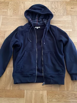 Svart hoodie från Burberry - Snygg svart hoodie från Burberry med dragkedja, två stora fickor framtill och klassisk huva med rutigt foder i grått, rött och svart. Diskret Burberry-logga broderad på bröstet. Perfekt för en avslappnad och stilren look.