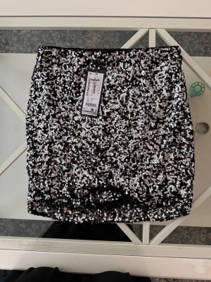 Svart paljettkjol från New Yorker - Säljer en svart kortkjol från New Yorker täckt med glittriga paljetter. Kjolen har en tajt passform och är perfekt för dig som vill sticka ut. Materialet är polyester och kjolen har en snygg, festlig look.