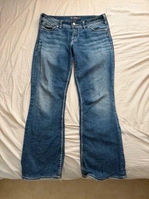 Blå bootcut jeans från Silver Jeans - Säljer ett par snygga blå bootcut jeans från Silver Jeans, modell Suki Surplus. Jeansen har klassisk femficksdesign, slitningar framtill och kontrastsömmar. Perfekta för dig som gillar en avslappnad men trendig look. W30 L34
