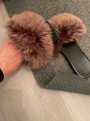Svarta tofflor med fluffig brun päls - Säljer ett par trendiga svarta tofflor med bred rem täckt av mjuk, fluffig brun fuskpäls. Snygg design som ger en lyxig känsla och passar perfekt för dig som vill ha något extra till hemmalooken. Platta sulor och öppen tå för maximal komfort.