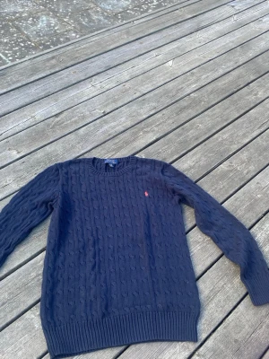 Mörkblå kabelstickad tröja Polo Ralph Lauren - Mörkblå kabelstickad tröja från Polo Ralph Lauren med rund halsringning och det klassiska röda broderade logot på bröstet. Tröjan har ribbade muddar vid ärmslut och nederkant, perfekt för en stilren och avslappnad look.