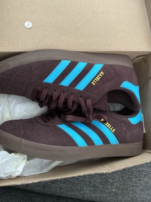 Adidas Gazelle brun/blå sneakers - Säljer ett par Adidas Gazelle sneakers i mocka med mörkbrun bas och ljusblåa klassiska tre ränder på sidorna.  Pris diskuteras , använd max 4 ggr