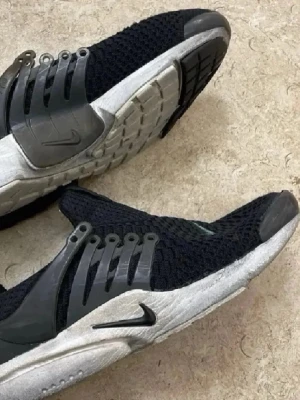 Nike svarta mesh sneakers - Säljer ett par svarta Nike sneakers med stickad mesh-överdel och grå plastdetaljer på sidorna. Skorna har en vit och svart sula med tydligt Nike-logga på sidan. Snygg och sportig design med låg profil och ventilerande material.