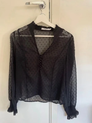 Svart transparent blus från NA-KD - Snygg svart transparent blus från NA-KD med diskreta prickar, volangdetaljer vid halsen och knappar framtill. Blusen har långa ärmar med resår och volang vid ärmslut. Perfekt för att lyfta vilken outfit som helst.