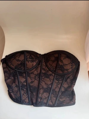Svart spets topp med bygel  - Snygg svart korsett med bygel och transparent spetsdetaljer. Axelbandslös modell med markerade sömmar och hakknäppning bak. Materialet är en kombination av mesh och spets som ger en lyxig känsla och snygg passform.
