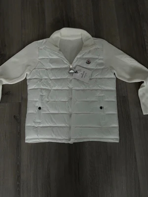 Vit moncler cardigan - Snygg vit dunväst från Moncler med quiltad front och två svarta knappar på fickorna. Västen har en hög ribbad krage och Moncler-logga på bröstet. Perfekt för lager-på-lager och ger en clean look till din outfit.
