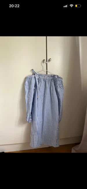 Blåvit randig offshoulder klänning - Säljer en blåvit randig kortklänning med offshoulder och långa puffärmar. Klänningen har resår vid ärmsluten och en knytning vid axeln för extra detalj. Perfekt för sommarens alla tillfällen!