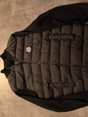 Svart dunväst från Moncler - Snygg svart dunväst från Moncler med quiltad framsida och stickade ärmar. Västen har två fickor med knappstängning och dragkedja framtill. Klassisk Moncler-logga på bröstet. Perfekt för lager-på-lager-stil.
