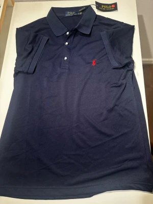 Marinblå Ralph Lauren pikétröja - Marinblå pikétröja från Polo Ralph Lauren med klassisk krage, tre knappar framtill och röd broderad logga på bröstet. Tröjan är helt ny och storlek S.