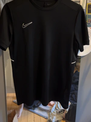 Svart Nike sporttröja - Snygg svart sporttröja från Nike med korta ärmar och vit logga på bröstet. Tillverkad i ett lätt och snabbtorkande funktionsmaterial som passar perfekt till träning eller match. Enkel och stilren design med små vita detaljer vid sidorna.
