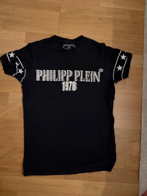 Svart Philipp Plein t-shirt med tryck - Snygg svart t-shirt från Philipp Plein med stort vitt tryck 'PHILIPP PLEIN 1978' på bröstet och stjärndetaljer på ärmarna. Klassisk passform och rund hals. Perfekt för dig som gillar streetwear och vill sticka ut.