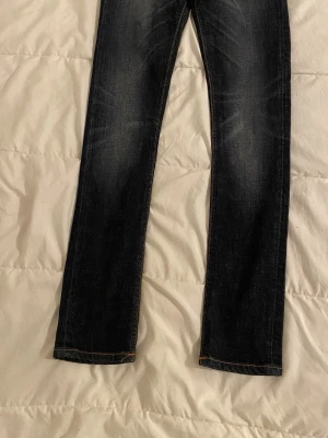 Mörkblå jeans från Nudie Jeans storlek W27, L30.  - Säljer ett par mörkblå jeans från Nudie Jeans med raka ben och snygga slitningar framtill. Jeansen har klassiska orange sömmar och bakfickor med Nudie Jeans signaturbrodyr. Perfekta för en avslappnad och stilren look.