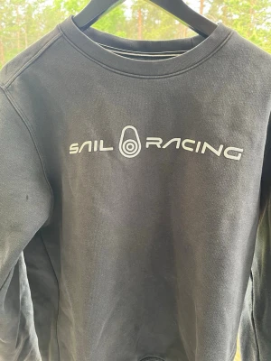 Svart sweatshirt från Sail Racing - Cool svart sweatshirt från Sail Racing med vit logga och tryck på bröstet samt en mindre logga i nacken. Klassisk rund halsringning och långa ärmar. Perfekt till jeans eller joggers för en avslappnad look. Kan gå ner i pris 