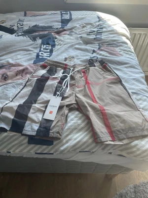 Bubber shorts med snörning - Snygga beige shorts med breda svarta och röda ränder. Shortsen har elastisk midja och vit snörning framtill för skön passform. Perfekta för sommaren och har en modern, avslappnad stil.