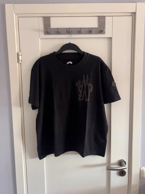 Svart Moncler t-shirt med tryck - Snygg svart t-shirt från Moncler med grafiskt tryck på bröstet och diskret logga på ärmen. Klassisk passform och rund halsringning, tillverkad i mjuk bomull. Perfekt för en stilren och avslappnad look.