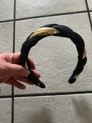 Flätat svart och guldigt diadem - Snyggt diadem med flätad design i svart tyg och guldigt material. Diademet har en bred form och detaljer i metall som ger en extra touch. Perfekt accessoar för att lyfta din look med lite extra bling.