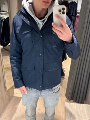 Mörkblå vindjacka med huva - Bondelid Jacka Mörkblå, använd 2 gånger! NYPRISET:1799kr.         Snygg mörkblå vindjacka med fyra fickor framtill och knappstängning. Jackan har en justerbar huva och är tillverkad i ett lätt, vattenavvisande material. Perfekt för blåsiga dagar och enkel att matcha med olika outfits.