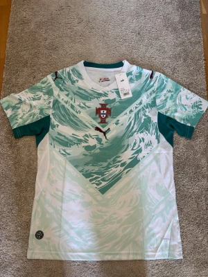 Portugal fotbollströja från Puma - Snygg Portugal fotbollströja från Puma med unikt mönster i mintgrönt och vitt. Tröjan har korta ärmar, rund hals och Portugal-emblem på bröstet. Perfekt för dig som älskar fotboll och vill sticka ut på planen eller läktaren.