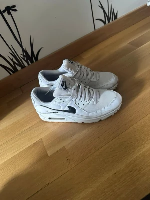 Nike Air Max 90 vita sneakers - Snygga vita Nike Air Max 90 sneakers med svart swoosh och detaljer. Skorna har en klassisk siluett med mesh och läder på ovandelen samt synlig Air-enhet i sulan. Perfekta för dig som gillar streetwear och sportig stil. Använt fåtal gånger, säljer pga att de e för små för mig