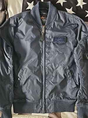 Mörkblå bomberjacka Alpha Industries - Snygg mörkblå bomberjacka från Alpha Industries med ribbade muddar, dragkedja framtill och två snedställda fickor med knapp. Jackan har en patch med logga på bröstet och klassisk ärmficka med dragkedja. Tillverkad i slitstarkt nylon material. Knapp i höger ficka har lossnat som syns på bild