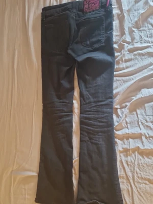 Svarta bootcut jeans från Miss Sixty - Snygga svarta jeans från Cheap Monday med bootcut passform och klassiska fem fickor. Jeansen har en mörk tvätt och en lila logotyppatch bak i midjan. Perfekta för dig som gillar en retroinspirerad look och vill ha något som sticker ut lite extra.