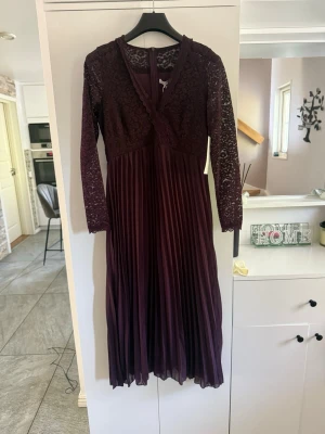 Vinröd långklänning med spetsdetaljer - Vinröd långklänning med plisserad kjol och överdel i spets. Klänningen har v-ringning och långa spetsärmar som ger en elegant look. Perfekt för dig som vill sticka ut med stilrena detaljer och en djup färg.