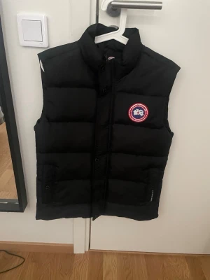 Svart dunväst från Canada Goose - Snygg svart dunväst från Canada Goose med hög krage och klassisk logotyp på bröstet. Västen har två framfickor med knappstängning och quiltad design som ger en puffig look. Perfekt för lager-på-lager och håller dig varm under kyliga dagar.
