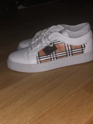 Burberry - Säljer ett par vita sneakers från Burberry med det klassiska rutiga mönstret i beige, svart, vitt och rött på sidan. Skorna har vita snören, vit sula och är tillverkade i slätt läder. Perfekta för dig som vill ha en stilren och trendig look.