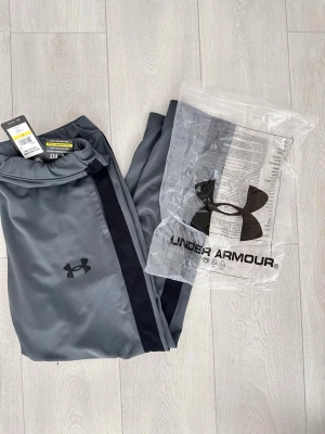 Grå träningsbyxor från Under Armour - Snygga grå träningsbyxor från Under Armour med svarta detaljer längs sidorna och logga på benet. Tillverkade i ett mjukt och stretchigt funktionsmaterial som passar perfekt till gymmet eller löprundan. Dragsko i midjan för bästa passform.