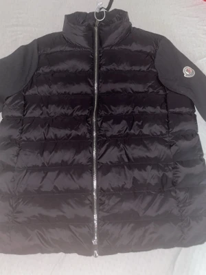 Svart dunväst från Moncler - Snygg svart dunväst från Moncler med quiltad design och hög krage. Västen har dragkedja framtill och ett Moncler-märke på axeln. Tillverkad i glansigt material som ger en exklusiv känsla. Perfekt för lager-på-lager och streetstyle. Priset går och pruta!
