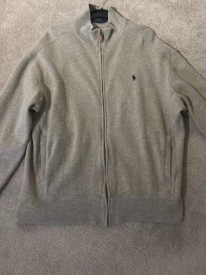 Grå ziptröja från Polo Ralph Lauren - Snygg grå ziptröja från Polo Ralph Lauren med hög krage och dragkedja hela vägen. Tröjan har diskret logga broderad på bröstet och två fickor framtill. Tillverkad i mjukt bomullsmaterial som är perfekt för lager-på-lager.