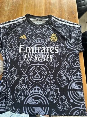 Real Madrid matchtröja Mbappé svart - Snygg svart Real Madrid matchtröja från Adidas med Mbappé och nummer 9 på ryggen. Tröjan har ett unikt mönster i grått, vita detaljer och klubbmärke på bröstet. Tillverkad i lätt och ventilerande material, perfekt för fotboll eller träning.