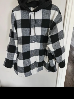 Svartvit rutig overshirt med huva - Overshirt i svartvitt rutigt mönster med tryckknappar och svart huva med dragsko. Tillverkad i ett mjukt, tjockare tyg som ger en avslappnad och trendig look. Perfekt för lager på lager och streetwear-stil.