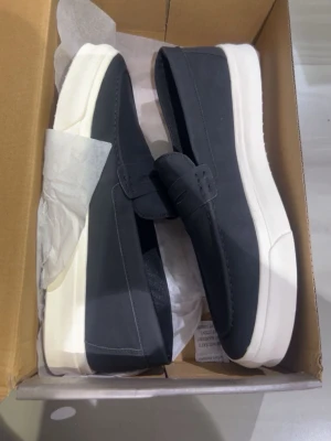 Svarta loafers med vit sula - Stilrena svarta loafers med bred vit sula och minimalistisk design. Skorna har en klassisk pennyloafers-detalj över vristen och är tillverkade i ett slätt material som ger en modern look. Perfekta för dig som gillar enkel och clean stil.