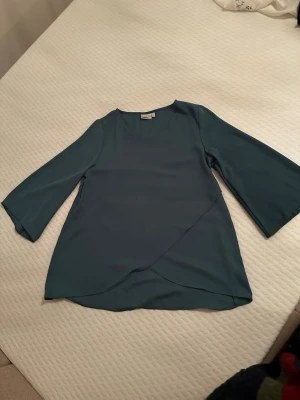 Grön omlottopp från Isolde - Snygg grön omlottopp från Isolde med trekvartsärm och v-ringning. Toppen har en elegant omlottdetalj framtill och är tillverkad i ett lätt, mjukt material som ger ett fint fall. Perfekt att styla med jeans eller kjol.