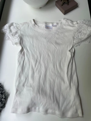 Vit topp med spetsärmar från Gina Tricot - Supersöt vit topp från Gina Tricot med ribbad struktur och korta ärmar i spets med broderade detaljer. Perfekt för dig som gillar en feminin och fräsch stil. Rund halsringning och mjukt material som känns skönt mot huden.