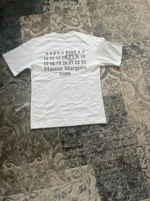 Maison Margiela vit t-shirt med tryck storlek S - Vit t-shirt från Maison Margiela med ikoniskt siffertryck och logga på ryggen. Klassisk rund hals och korta ärmar. Tillverkad i mjuk bomull för en clean och stilren look. Perfekt för dig som gillar minimalistisk design med en twist. T-shirten är helt ny och aldrig buren och har storlek S