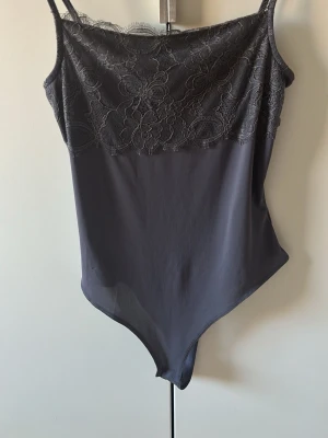 Mörkgrå spetsbody med smala band - Snygg mörkgrå body med smala axelband och spetsdetaljer över bysten. Överdelen har ett blommigt spetsmönster och resten är i slät, stretchig textil. Perfekt för att skapa en trendig lager-på-lager-look.