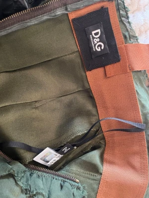 Olivgrön omlottklänning från D&G - Perfekt för en soirée eller en fin middag🍸🥂 Den här snygga olivgrön omlottklänning från D&G i glansigt material är en must-have i varje fashionistas garderob💗. Klänningen har djup v-ringning, breda axelband och markerad midja med brun detalj. Kort modell som ger en elegant siluett, perfekt för dig som vill sticka ut. 